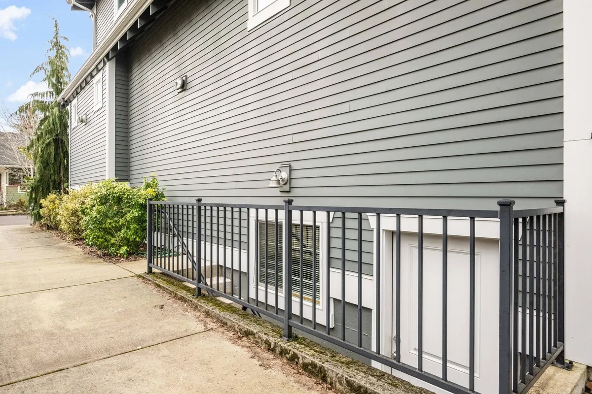 An exterior side view of the house surrounded by greenery and a railing, evoking a tranquil mood. The scene suggests a gentle transition from the outside world into a secluded, peaceful space, reinforcing the idea of a tucked-away haven.