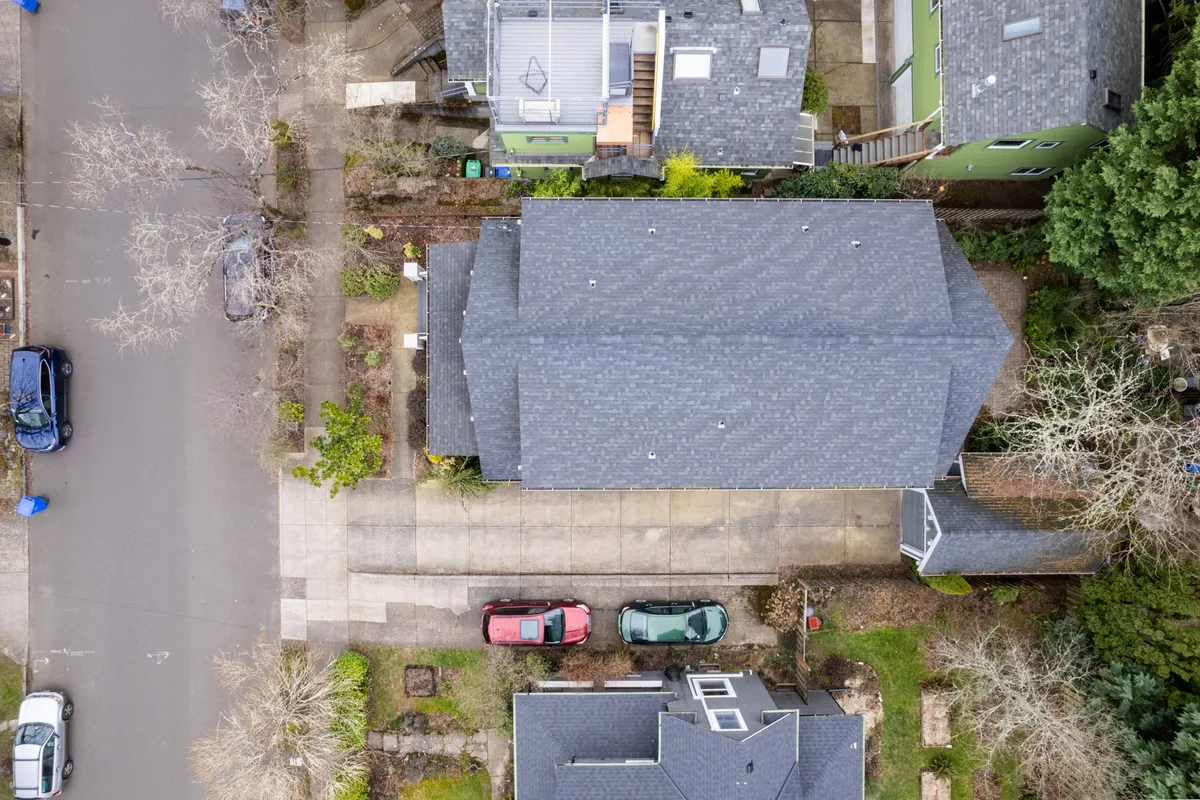 A calm, tree-lined residential street in Sellwood featuring charming rooftops, green yards, and several cars parked in front. The scene shows a welcoming neighborhood atmosphere, perfect for strolling and accessing local shops or cafes. The greenery and driveways suggest a peaceful yet accessible location, ideal for visitors interested in exploring on foot.