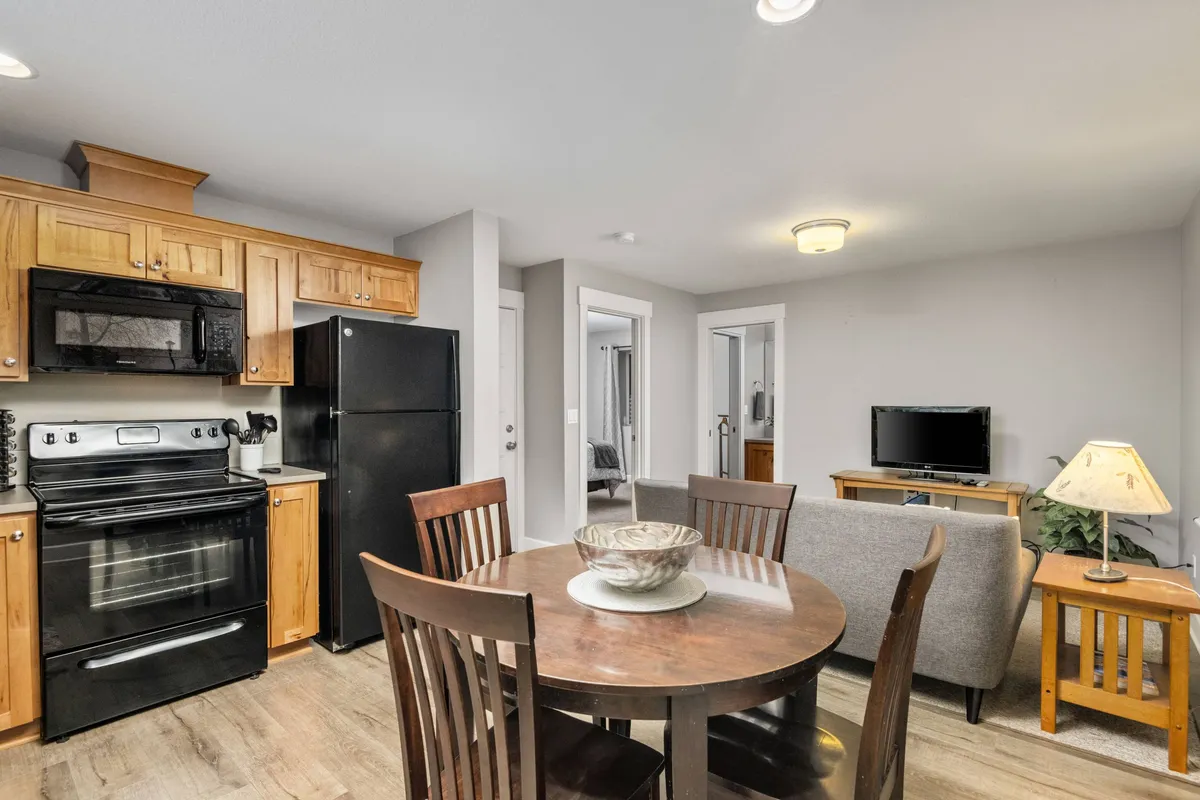 This image showcases an open interior where the kitchen, dining table, and a comfortable seating area with a television are seamlessly integrated. The space features cozy furnishings, ample natural light from windows, and is arranged for both socializing and relaxation.